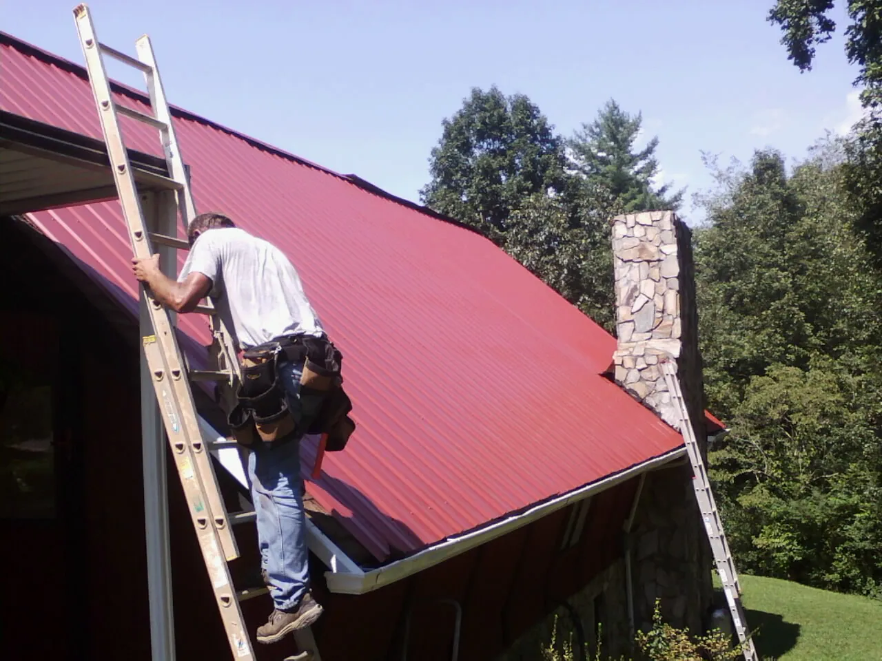 Roofer on a ladder beside a red metal roof and stone chimney — Soffit & Fascia Repair by Roofing Brothers in Rockfall